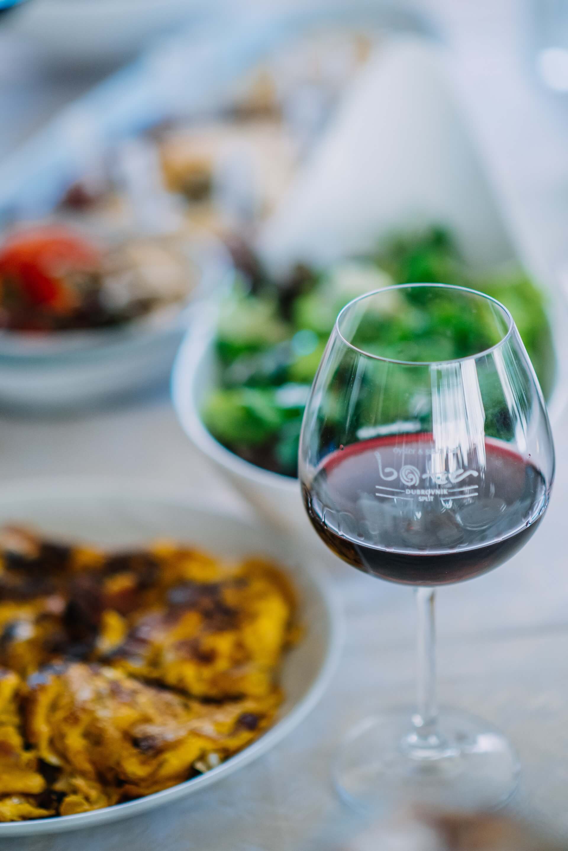 A glass of red wine with grilled food in the background.
