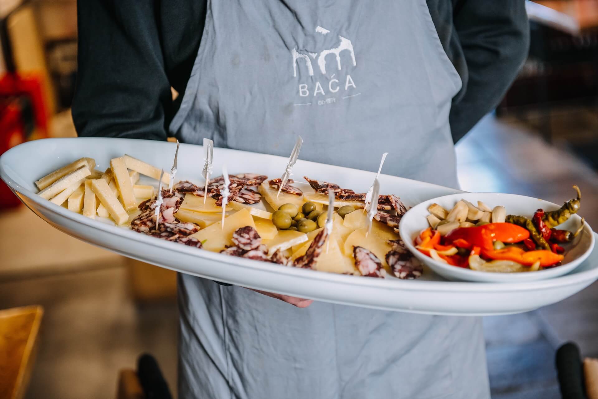 A platter of Pelješac prosciutto and paški sir, with marinated peppers and olives, carried by a server in a Baća apron.